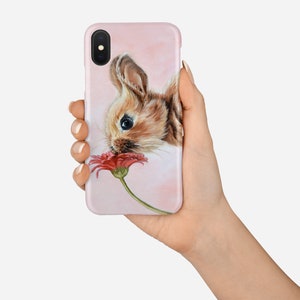 May include: A pink iPhone case featuring a painting of a brown bunny rabbit smelling a red flower.