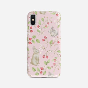 May include: A pink phone case with a repeating pattern of white flowers, green leaves, and red strawberries. Two small gray rabbits are featured in the pattern.