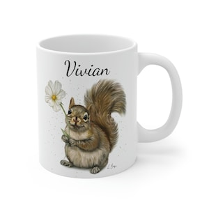 Peut inclure: Une tasse en céramique blanche avec un écureuil brun tenant une fleur blanche. La tasse est personnalisée avec le nom "Vivian".