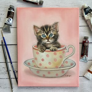 Puede incluir: Una pintura de un gatito atigrado marr&oacute;n y negro sentado en una taza de t&eacute; rosa con lunares blancos. El gatito tiene ojos azules y mira al espectador. La taza de t&eacute; est&aacute; sobre un platillo blanco con lunares rosas. El fondo es rosa claro.