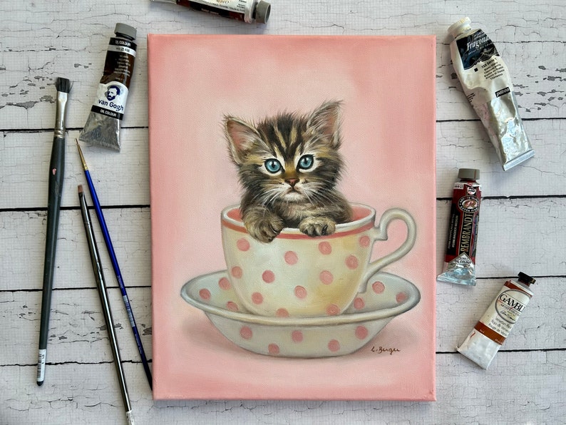Puede incluir: Una pintura de un gatito atigrado marr&oacute;n y negro sentado en una taza de t&eacute; rosa con lunares blancos. El gatito tiene ojos azules y mira al espectador. La taza de t&eacute; est&aacute; sobre un platillo blanco con lunares rosas. El fondo es rosa claro.