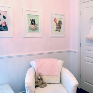 Puede incluir: Una habitaci&oacute;n de beb&eacute; rosa y blanca con un sill&oacute;n blanco, una manta rosa y blanca y un conejito de peluche. Hay tres cuadros enmarcados en la pared con im&aacute;genes de un panda, un gatito y una ardilla.