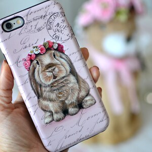 May include: A pink phone case featuring a detailed illustration of a brown bunny wearing a floral crown. The case has a vintage aesthetic with script and a stamp design. The bunny is the focal point, with soft fur and a gentle expression.