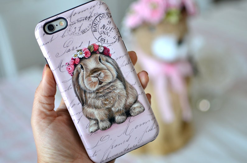 May include: A pink phone case featuring a detailed illustration of a brown bunny wearing a floral crown. The case has a vintage aesthetic with script and a stamp design. The bunny is the focal point, with soft fur and a gentle expression.