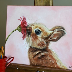Puede incluir: Una pintura de un conejo marr&oacute;n oliendo una flor roja sobre un fondo rosa. El conejo tiene un ojo negro y un pelaje marr&oacute;n y esponjoso. La pintura est&aacute; firmada "L. Bergen" en la esquina inferior derecha.