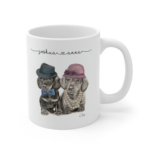 May include: White ceramic mug with a black and white illustration of two dachshunds wearing hats. The dachshund on the left is wearing a blue hat and bow tie. The dachshund on the right is wearing a pink hat and a pearl necklace. The text "joshua ♥ anna" is written above the dachshunds.