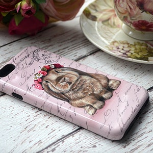 May include: A pink phone case featuring a sleeping bunny wearing a floral crown. The case has a vintage design with script writing and a postage stamp graphic. The bunny is brown with detailed fur.