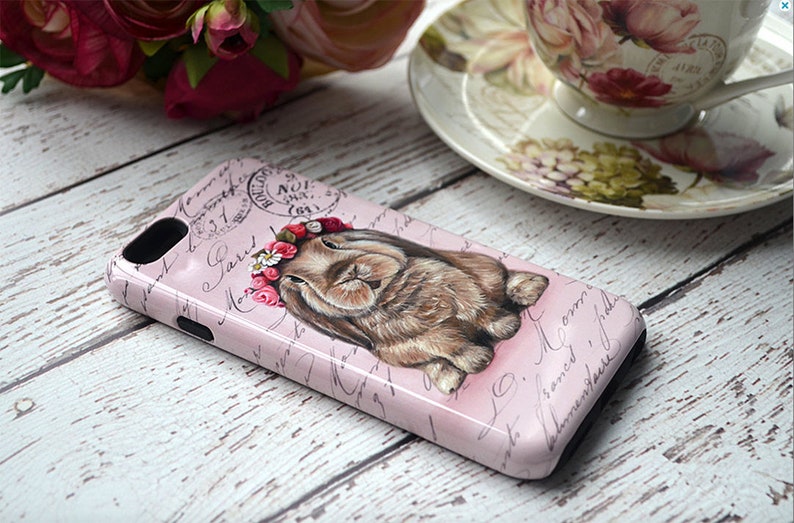 May include: A pink phone case featuring a sleeping bunny wearing a floral crown. The case has a vintage design with script writing and a postage stamp graphic. The bunny is brown with detailed fur.