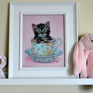 Puede incluir: Una pintura enmarcada de un gatito con ojos azules sentado en una taza de t&eacute; de lunares rosa y blanco. La pintura est&aacute; en un estante blanco con un juguete de peluche de conejo rosa a la derecha.