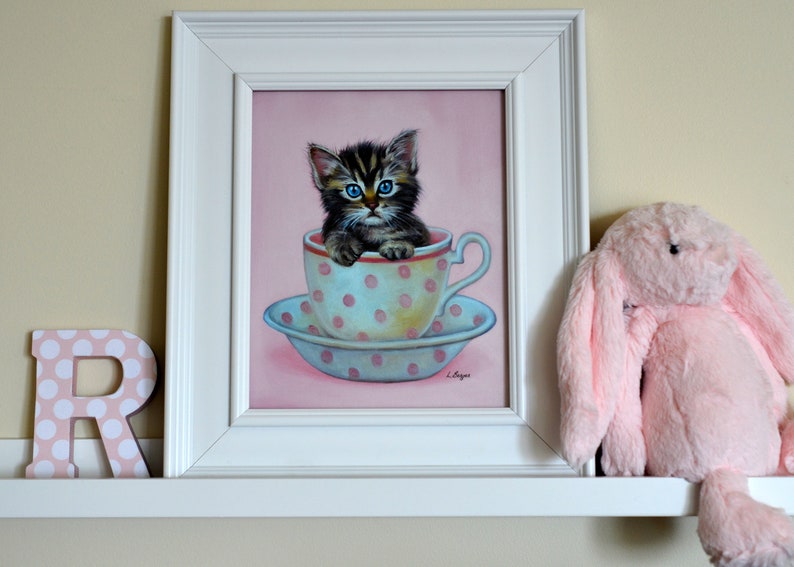 Puede incluir: Una pintura enmarcada de un gatito con ojos azules sentado en una taza de t&eacute; de lunares rosa y blanco. La pintura est&aacute; en un estante blanco con un juguete de peluche de conejo rosa a la derecha.