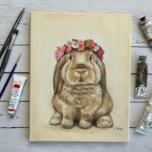 May include: An original painting of a brown rabbit wearing a floral crown. The artwork is on a canvas, with paint tubes and brushes arranged around it. The rabbit has long ears and a fluffy coat. The painting is signed by the artist.