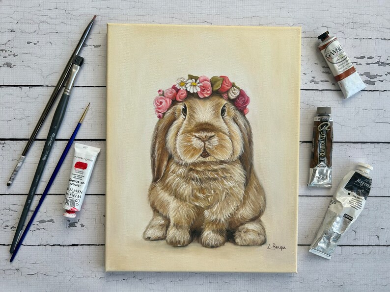May include: An original painting of a brown rabbit wearing a floral crown. The artwork is on a canvas, with paint tubes and brushes arranged around it. The rabbit has long ears and a fluffy coat. The painting is signed by the artist.