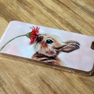 Puede incluir: Una funda de tel&eacute;fono transparente con un dise&ntilde;o pintado de un conejo marr&oacute;n oliendo una flor roja.