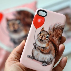 May include: A pink phone case with a squirrel holding a red balloon. The squirrel is brown and white with a bushy tail. The background is a light pink.