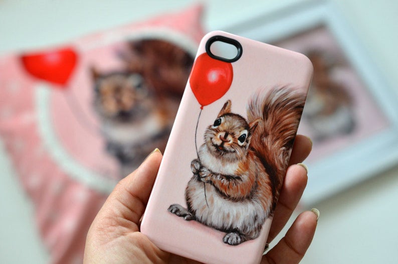 May include: A pink phone case with a squirrel holding a red balloon. The squirrel is brown and white with a bushy tail. The background is a light pink.