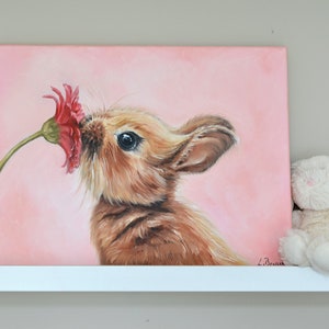 May include: A painting of a brown bunny rabbit sniffing a pink flower on a pink background.