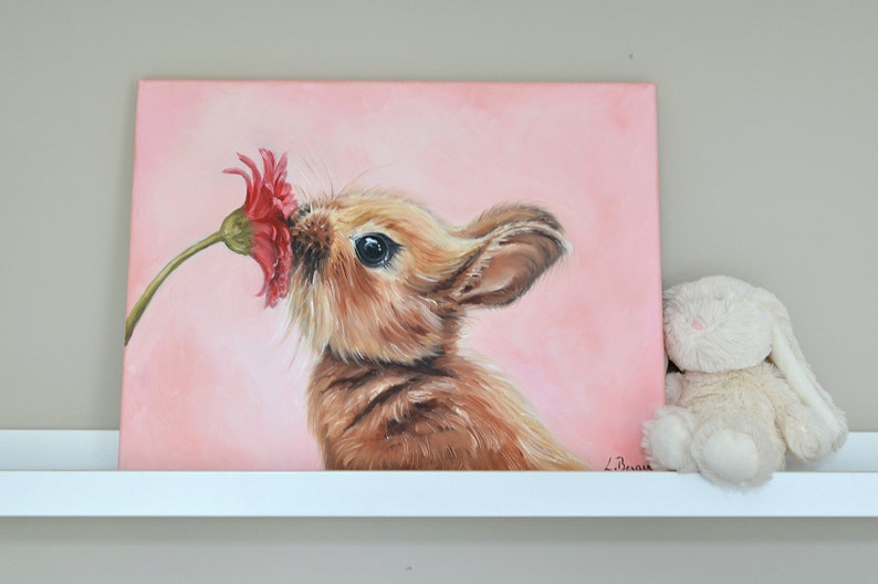 May include: A painting of a brown bunny rabbit sniffing a pink flower on a pink background.