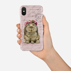May include: A phone case featuring a brown rabbit wearing a floral crown. The case has a pink background with faded script and postage stamps. The phone case is held in a hand.