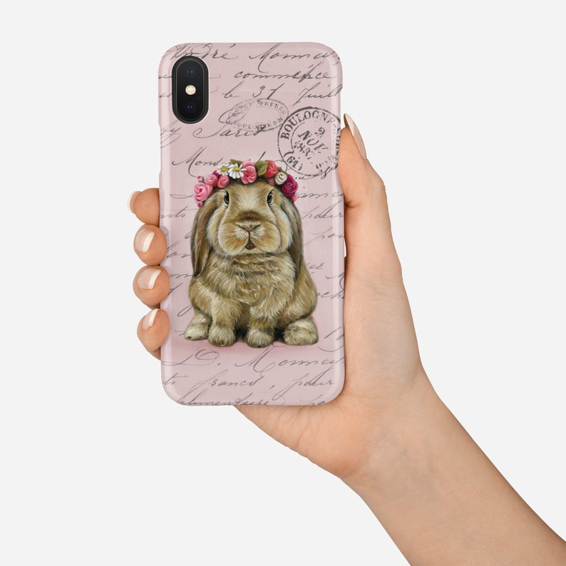 May include: A phone case featuring a brown rabbit wearing a floral crown. The case has a pink background with faded script and postage stamps. The phone case is held in a hand.
