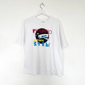 Op de afbeelding: Wit t-shirt met een grafisch ontwerp met de woorden "РЕГУЛЯРНО ЧИСТИТЕ ЗУБЫ" in rood, blauw en geel. Het ontwerp bevat een afbeelding van een tube tandpasta en een tandenborstel. Het t-shirt hangt aan een witte hanger.