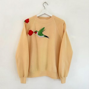 May include: A light yellow sweatshirt with a crew neck and long sleeves. The sweatshirt features an embroidered hummingbird with a red flower in its beak. Another red flower is embroidered on the shoulder. The sweatshirt is hanging on a white hanger.