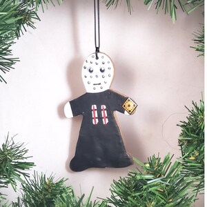 May include: A hand-painted gingerbread ornament shaped like a person wearing a black robe and a white mask with black dots. The ornament has a red and white striped pattern on the robe and a yellow and black patch on the right arm.