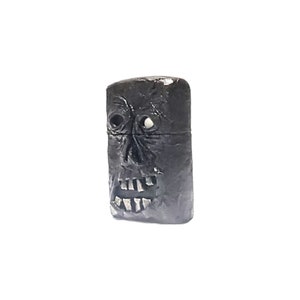 May include: A rectangular black lighter featuring a sculpted, textured face on the front surface. The design includes deep-set eye sockets, a prominent nose, and an open mouth revealing individual teeth. The lighter has a horizontal seam across the middle.