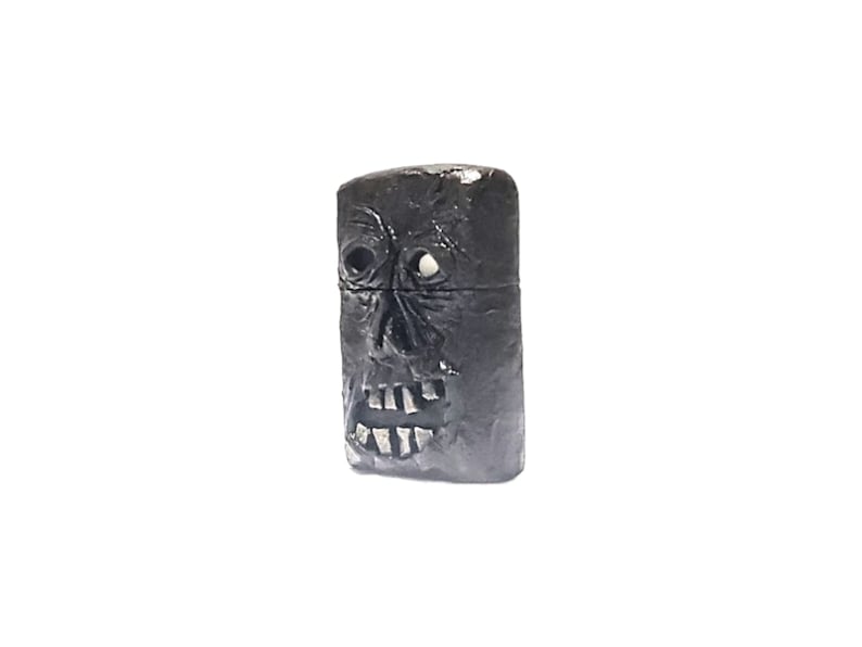 May include: A rectangular black lighter featuring a sculpted, textured face on the front surface. The design includes deep-set eye sockets, a prominent nose, and an open mouth revealing individual teeth. The lighter has a horizontal seam across the middle.