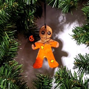 May include: Orange and brown gingerbread man ornament with a stitched face and a pumpkin-shaped lollipop. The ornament is hanging from a black string.