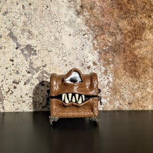 May include: A brown, monster-themed box with a hinged lid and silver-colored feet. The box features a large, toothy grin and a single eye. The box is likely a decorative item or a unique storage container.
