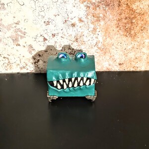 May include: A teal-colored, rectangular box with a cartoon alligator design. The alligator has white teeth, blue eyes, and silver feet. The box is sitting on a black surface.