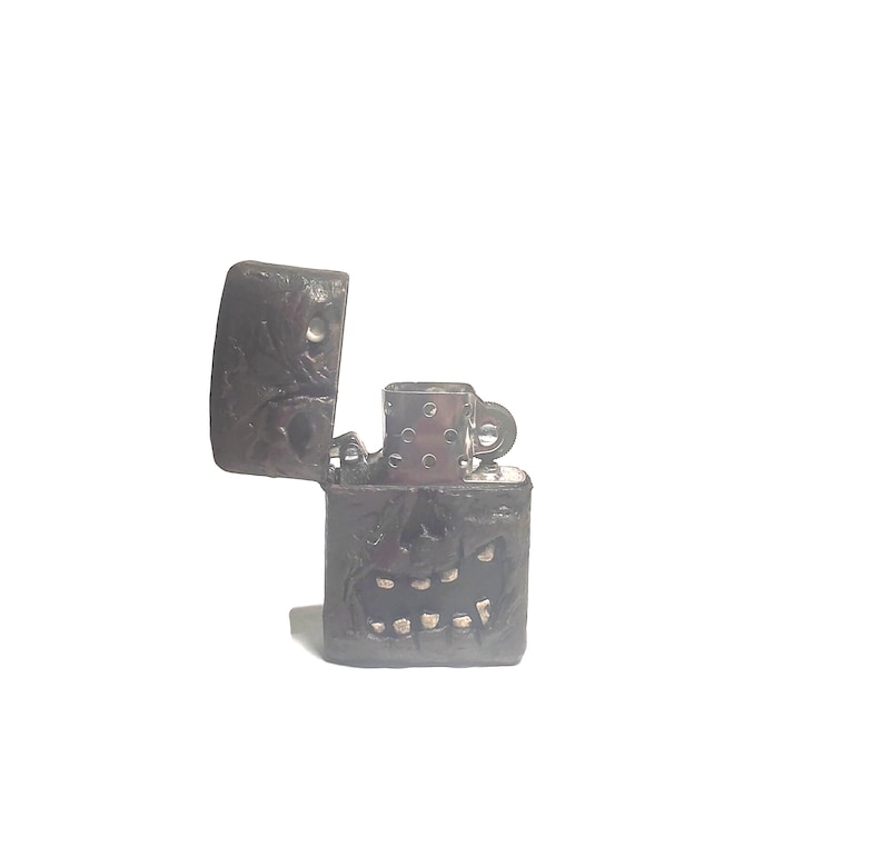 May include: A dark brown, textured metal lighter with an open lid, featuring a sculpted, skull-like face on the front with visible teeth and hollow eye sockets. The internal silver-colored metal insert is partially visible within the casing.