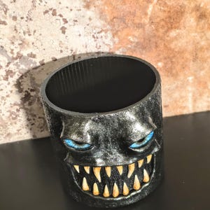 May include: A cylindrical black container featuring a sculpted monster face with textured skin, piercing blue eyes, and a wide grin displaying sharp, pointed yellow teeth. The interior of the vessel is smooth and black, contrasting with the exterior.