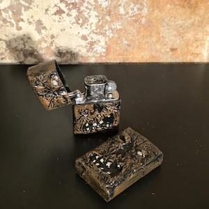 May include: A metal flip-top lighter with a textured, dark bronze-colored exterior featuring a raised, rugged pattern and small white accents. The lighter is shown in two pieces, with the hinged lid open and the internal silver-colored mechanism visible.