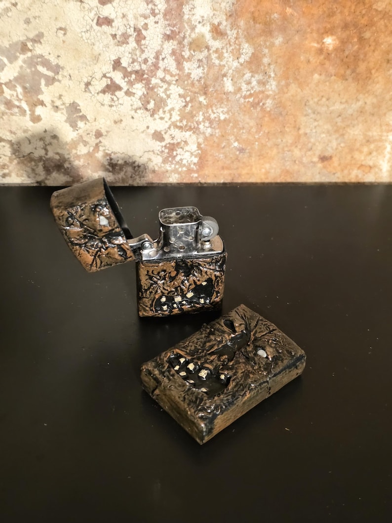 May include: A metal flip-top lighter with a textured, dark bronze-colored exterior featuring a raised, rugged pattern and small white accents. The lighter is shown in two pieces, with the hinged lid open and the internal silver-colored mechanism visible.