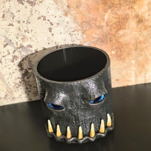 May include: A cylindrical black container with a textured surface, featuring two recessed blue eyes and a row of pointed, off-white teeth along the bottom edge. The interior of the container is smooth and black, with a wide, open top opening.