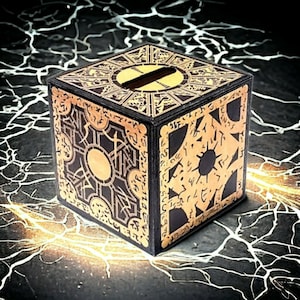 May include: A black wooden box with a gold intricate design. The box has a small opening on the top and is a replica of the iconic puzzle box from the movie "Saw".