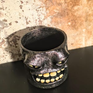 May include: A cylindrical container with a dark, metallic, textured finish featuring a sculpted face with narrow, gold-toned eyes and a wide, grinning mouth filled with prominent, off-white teeth. The object has a hollow interior and a dark rim.