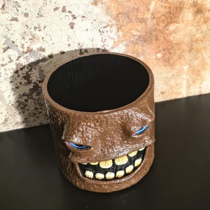 May include: A cylindrical brown planter with a textured surface featuring a sculpted face. The face has two narrow blue eyes and a wide, open mouth displaying a row of off-white teeth. The interior of the container is smooth and black.