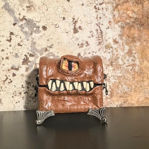 May include: A brown, rectangular box with a monster face design. The box features a large, yellow eye with red details, a wide mouth with white teeth, and decorative silver feet. The box is on a dark surface against a textured background.