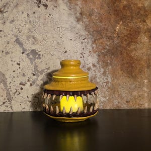 May include: A yellow jar with a lid, featuring a row of sharp, tooth-like protrusions around the middle. The jar has a glowing yellow light inside, creating a spooky effect. The jar is sitting on a dark surface.