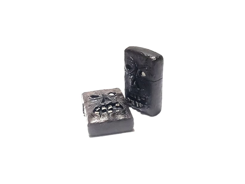 May include: Two rectangular black lighters featuring a sculpted, three-dimensional face on the front surface. Each lighter displays a detailed skull-like visage with hollow eye sockets and a bared-teeth mouth, finished in a dark, matte texture.
