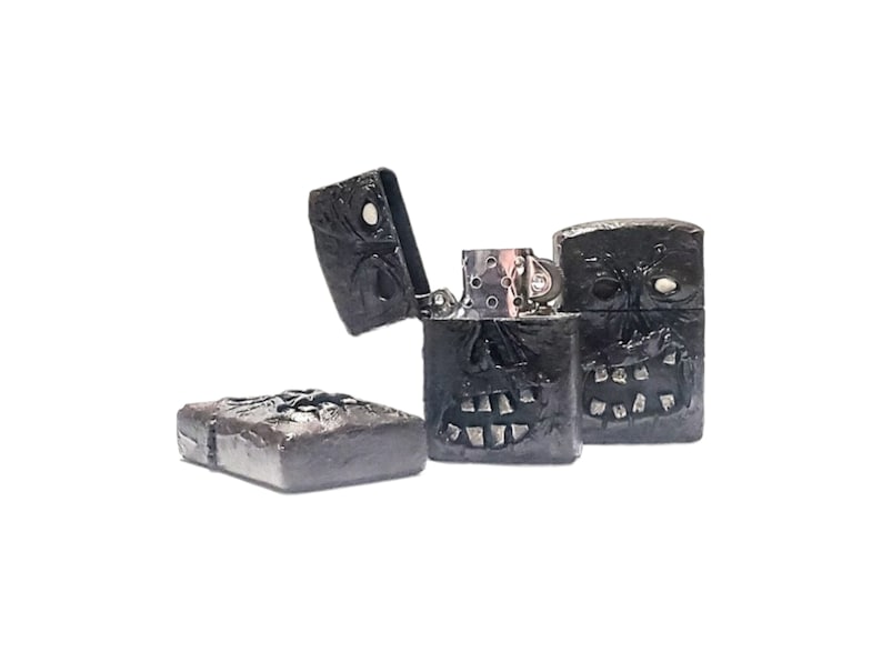 May include: Three black metal lighters with a textured, sculpted surface resembling a skull face. One lighter is open, revealing the silver-colored internal mechanism, while the other two are closed, showing the detailed relief of teeth and eyes.