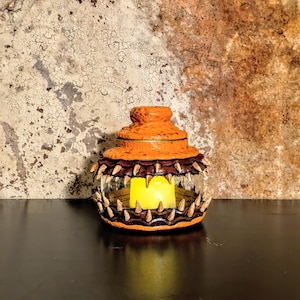 May include: An orange and brown decorative lantern with a glass center and a yellow candle inside. The lantern has a textured, spiky design around the edges, resembling teeth. The background is a textured wall.