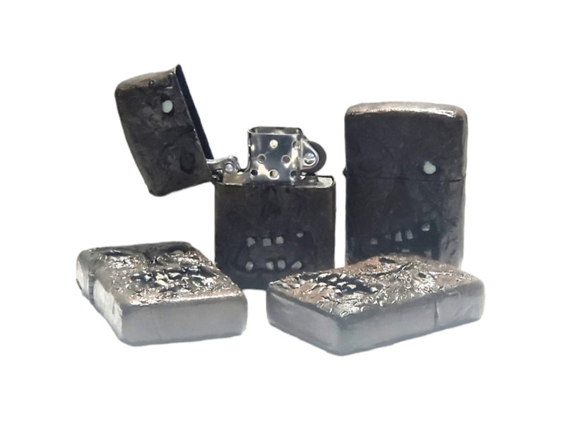 May include: A collection of four rectangular metal lighters with a dark, textured finish resembling a skull-like face. One lighter is open, revealing the silver-colored internal mechanism, while the others are closed and display the same relief design.