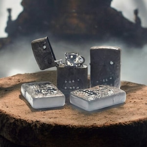 May include: Four rectangular metal lighters with dark, textured surfaces featuring skull-like motifs. One lighter is open, revealing the internal silver-toned mechanism with a chimney and flint wheel. Two lighters are lying flat, while two stand upright.