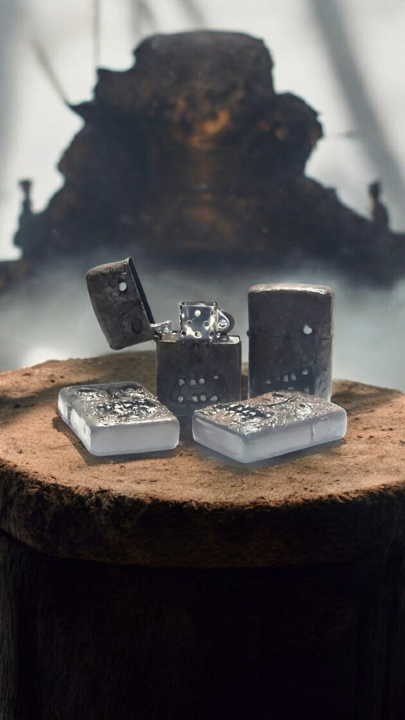May include: Four rectangular metal lighters with dark, textured surfaces featuring skull-like motifs. One lighter is open, revealing the internal silver-toned mechanism with a chimney and flint wheel. Two lighters are lying flat, while two stand upright.