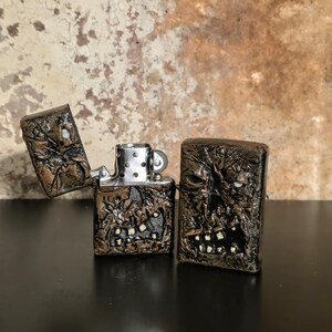 May include: Two rectangular metal lighters with a dark, textured, sculpted finish resembling a weathered, monstrous face with visible teeth. One lighter is open, revealing the silver-toned internal mechanism, while the other is closed.