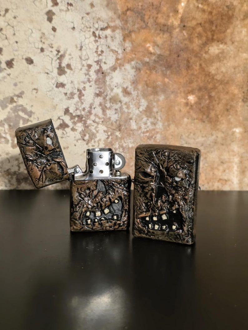 May include: Two rectangular metal lighters with a dark, textured, sculpted finish resembling a weathered, monstrous face with visible teeth. One lighter is open, revealing the silver-toned internal mechanism, while the other is closed.