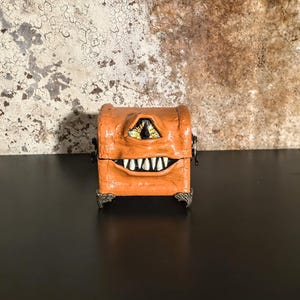 May include: Orange, handcrafted treasure chest with a monster face design. The chest features a large, yellow eye, a wide grin with white teeth, and decorative metal feet. The chest is a unique, whimsical decorative item.